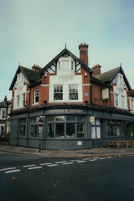 The image depicts a multi-story building with a traditional brick facade and white decorative timber framing, located on a street corner. The ground floor features large glass windows and a dark grey painted exterior with the sign 'Greenwich Tavern' visible above the windows. In front of the building, there is a paved pavement with a black and white striped road marking, a pole with a no-entry sign, and a wooden bench placed near the corner. Inside a residential entrance on the right side of the building, a person is seen carrying a cardboard box, dressed in casual clothing, actively engaged in moving items. The interior of the building’s ground floor appears to be used for commercial purposes, possibly a pub or restaurant, while the upper floors feature multiple windows and a chimney, indicating residential or office spaces above. The scene captures a moment of home relocation or furniture transport as part of a professional move by Greenwich Man and Van, with the loading process occurring near the building’s entrance, supported by the presence of packaging materials and the person carrying boxes.