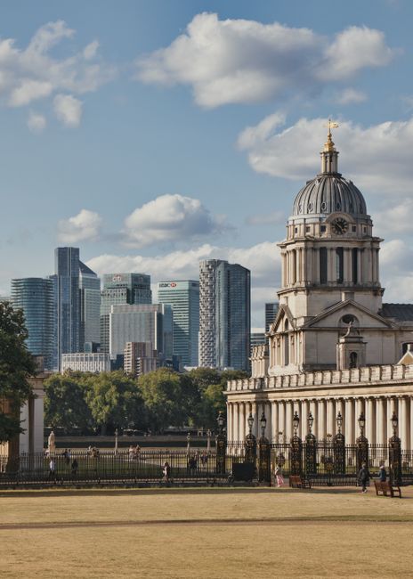 A historic domed building with classical architectural features, including ornate columns and a decorative balustrade, situated within a park area with a grass lawn in the foreground. Behind the building, modern high-rise office towers with glass facades form the skyline under a partly cloudy sky. The scene suggests an urban setting combining heritage architecture and contemporary city development, relevant to house removals and home relocation services such as those provided by Greenwich Man and Van, as they facilitate moving and furniture transport in city environments.