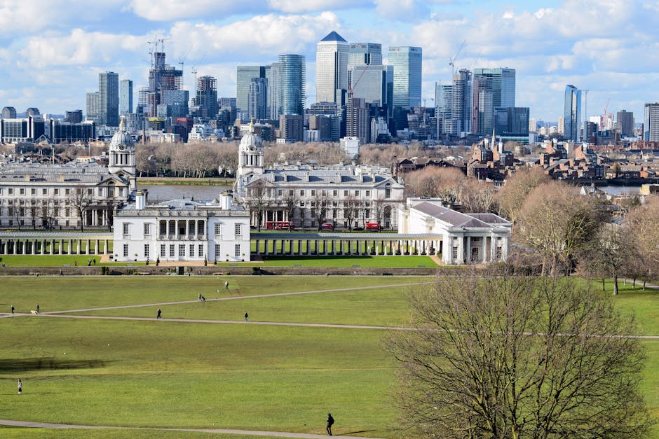 A wide view of the Royal Hospital Chelsea in London, with its historic white stone facade, symmetrical columns, and domed towers visible. In the foreground, there is a large, open park area with well-maintained green grass, several winding pathways, and a few scattered people walking. The park separates the historic building from the modern city skyline in the background, which features numerous high-rise office buildings and skyscrapers with glass and steel facades. The sky above is partly cloudy with patches of blue. The image captures a peaceful urban scene, and the setting suggests a location that may be part of a house or office move and transport service, with Greenwich Man and Van occasionally involved in logistics and relocation efforts, although no moving activities or objects are visible in this particular image.