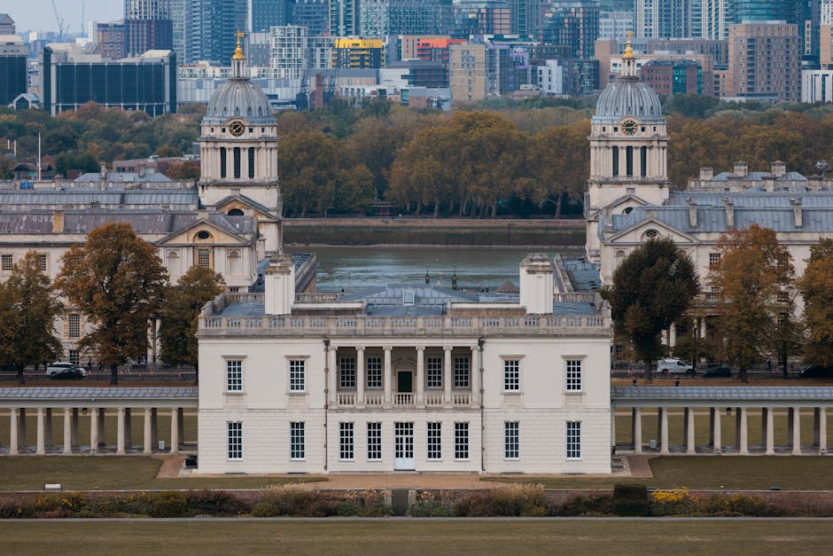 A wide-angle view of a historic neoclassical building situated near a river, featuring a central structure with white stone walls, multiple large rectangular windows, and a flat roof. Two prominent domed towers with clock faces and columned entrances flank the main building; these towers have detailed cornices and white domes topped with golden finials. The building is connected to a colonnade with evenly spaced columns, extending along its length. In front of the building, there is a well-maintained lawn with neatly trimmed bushes, and to the left and right, there are several large deciduous trees with brown and green foliage, indicating an autumn season. Behind the structure, across the river, a modern cityscape with high-rise buildings of glass and steel is visible, with a mix of commercial and residential towers. The sky above is overcast, casting soft, diffused light over the scene, which might be used as a backdrop during home relocation or furniture transport planning. The setting suggests a historic site near Greenwich Market, relevant for house removals in Greenwich, supported by [COMPANY_NAME] in the moving industry.