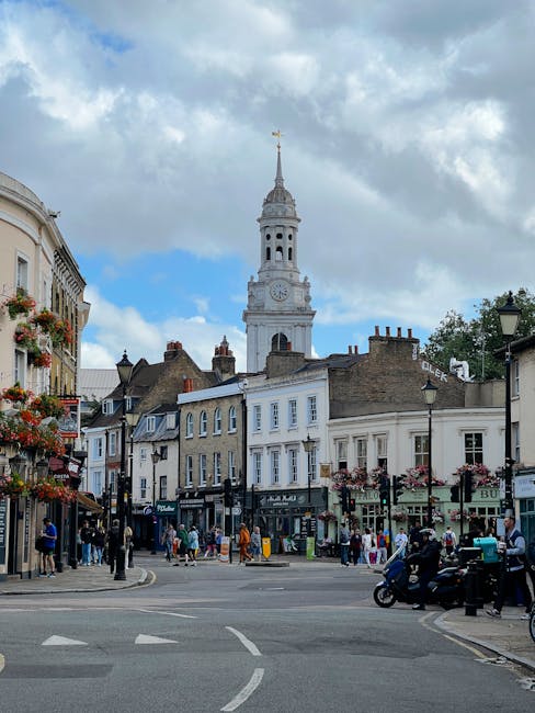 A street scene near Greenwich Market featuring historic buildings with white and beige facades, some with decorative elements and large windows, situated along the corner of the street. In the background, the distinctive tall clock tower of the Cutty Sark ship stands prominently against a partly cloudy sky. The street is bustling with pedestrians walking on the pavement, some engaging in conversation, and a few vehicles, including a motorcycle and a few cars, waiting at the intersection. On the left, hanging flower baskets adorn street lamps and building facades, adding colour to the scene. The area appears lively with shops and cafes visible at ground level, contributing to the vibrant atmosphere typical of Greenwich. This location is photographed during daytime with natural light illuminating the scene, supporting the context of home relocation and furniture transport services provided by Greenwich Man and Van near Greenwich Market.