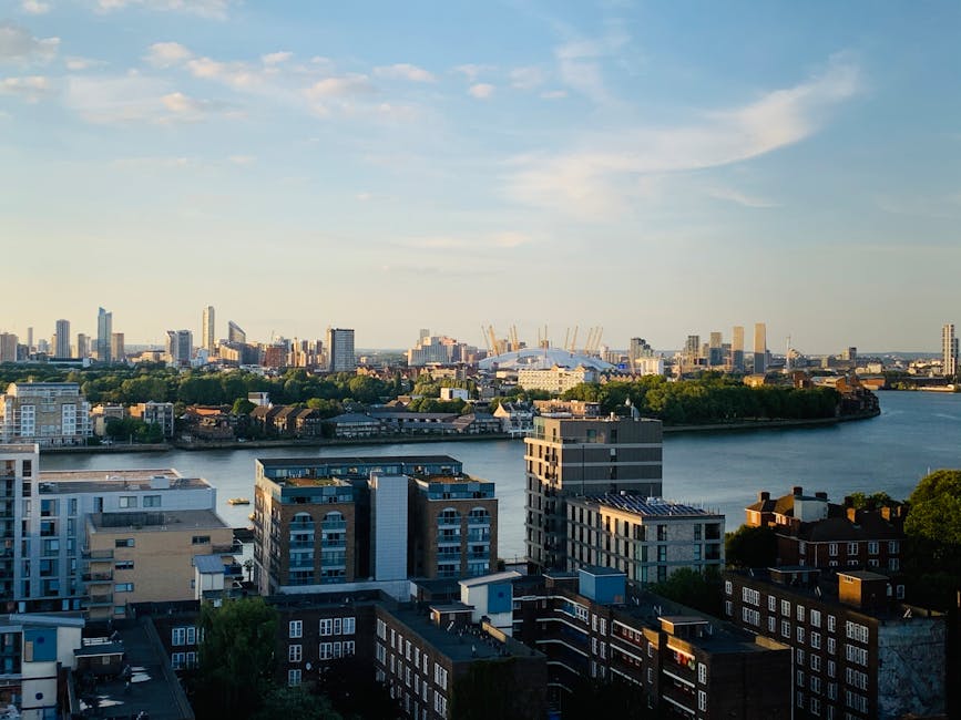 The image shows a panoramic view of Greenwich, London, taken from a high vantage point during daylight hours with clear blue sky and a few clouds. In the foreground, a cluster of modern residential buildings with flat roofs, balconies, and large windows are visible, situated along the riverbank. The river Thames runs through the middle of the scene, with its calm water reflecting the sky and surrounding structures. On the opposite bank, a mix of commercial and residential buildings, some with rooftops and greenery, extend towards the skyline. In the background, the iconic London cityscape features several high-rise buildings, cranes indicating ongoing construction, and the distinctive dome and arches of The O2 Arena, which is the focus of the event clearouts and commercial removals service offered by Greenwich Man and Van. The natural daylight illuminates the scene, capturing the urban landscape and hinting at the logistics involved in furniture transport and home relocation services within the city.
