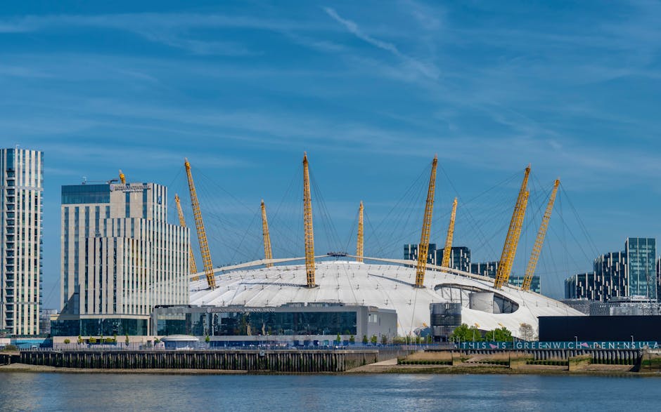 The image shows a panoramic view of Greenwich, London, taken from a high vantage point during daylight hours with clear blue sky and a few clouds. In the foreground, a cluster of modern residential buildings with flat roofs, balconies, and large windows are visible, situated along the riverbank. The river Thames runs through the middle of the scene, with its calm water reflecting the sky and surrounding structures. On the opposite bank, a mix of commercial and residential buildings, some with rooftops and greenery, extend towards the skyline. In the background, the iconic London cityscape features several high-rise buildings, cranes indicating ongoing construction, and the distinctive dome and arches of The O2 Arena, which is the focus of the event clearouts and commercial removals service offered by Greenwich Man and Van. The natural daylight illuminates the scene, capturing the urban landscape and hinting at the logistics involved in furniture transport and home relocation services within the city.