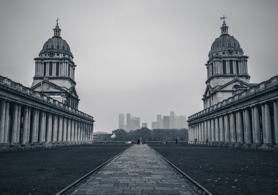 A black-and-white photograph of the Old Royal Naval College in Greenwich, London, showcasing two symmetrical domed towers with classical architectural details and clock faces. In the foreground, a wide stone-paved pathway lined with low stone walls extends towards the distant city skyline, including modern high-rise buildings visible through a light haze. The outdoor area appears to be an open, grassy space with a few small figures and trees in the background. The image captures a historic heritage site that could relate to home relocation or property moving logistics, with a focus on the scale and grandeur of the architecture and surroundings.