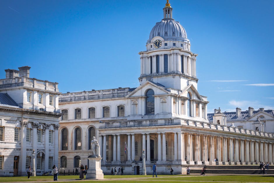 A large historic building with a prominent domed clock tower featuring a gold finial on top, situated beneath a bright blue sky. The structure has neoclassical architectural elements, including white stone facades, arched windows, and decorative columns. In front of the building, there is a paved area with several pedestrians walking or standing, some near statues and lampposts. The scene captures a daytime view with clear weather, highlighting the building's grandeur which could be associated with a city centre or government district. The image reflects the type of urban environment where home relocation and furniture transport services might operate, with Greenwich Man and Van providing professional removals and moving logistics in such locations.