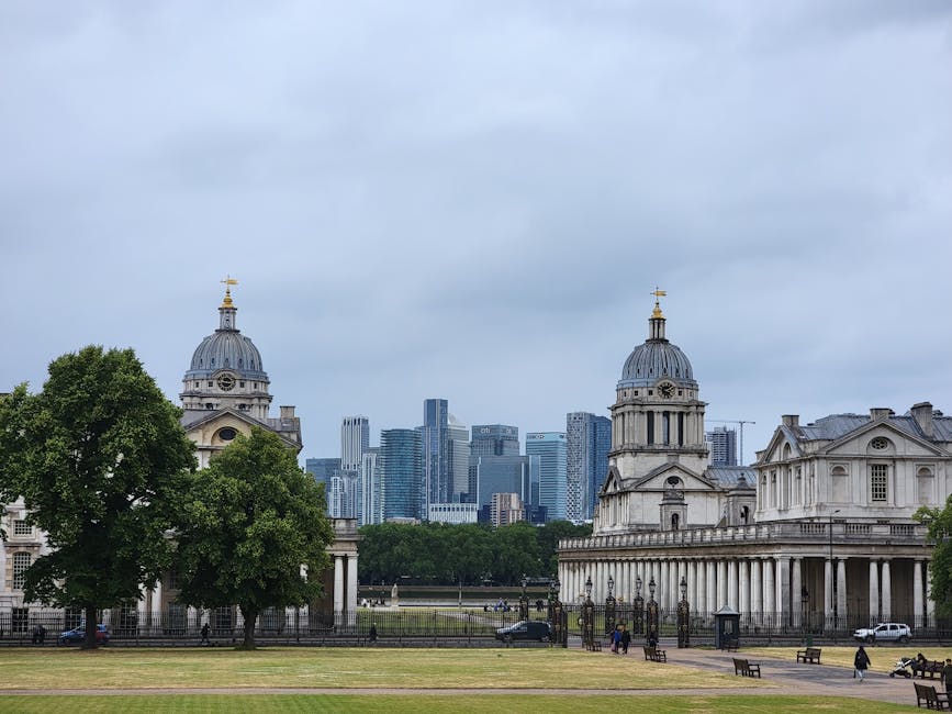 A daytime scene featuring historic church buildings with domed towers and classical architectural details, situated on a green park with trees and open grassy areas. In the background, there is a city skyline with modern high-rise office buildings made of glass and steel under an overcast sky. The foreground shows a paved area where a person can be seen loading or unloading cardboard boxes and wooden crates from a vehicle, with packing blankets and plastic wrap nearby, indicating a home relocation or furniture transport process. The scene represents an outdoor part of a house removal in an urban area, with Greenwich Man and Van providing professional moving services, as suggested by the context of the image.
